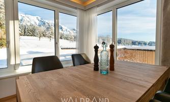 A bright dining room with a large wooden table and modern chairs. Through the windows, a snowy landscape with mountains can be seen.