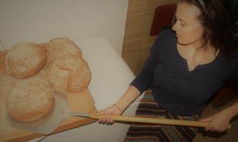 A woman sits next to fresh bread that is on a wooden board. She holds a baking tool in her hand and seems to be looking at the bread.
