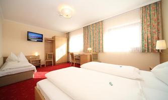 A bright hotel room with two single beds and an additional bed. There is a television, a desk, and windows with curtains.