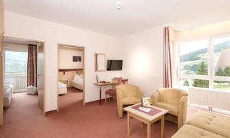A bright and spacious hotel room with a living area, sofas, and a table. From the windows, there is a view of the mountains.