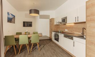 A modern kitchen with a dining table and green chairs. The walls are light, and a wooden paneling creates a cozy atmosphere.
