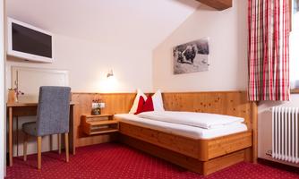 A cozy room with a single bed, desk, and television. The walls are made of wood, and there is a large window with red curtains.