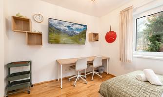 A modern bedroom with a bed, a desk, and two chairs. A TV is mounted on the wall, and there are large windows with a view to the outdoors.