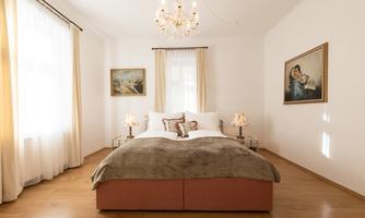 A bright bedroom with a large bed and elegant bedding. The walls are decorated with artwork and there are cozy nightstands with lamps.