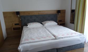 A cozy bedroom with a large double bed and elegant wooden design. The room is bright and welcoming, featuring a mirror and window curtains in soft colors.