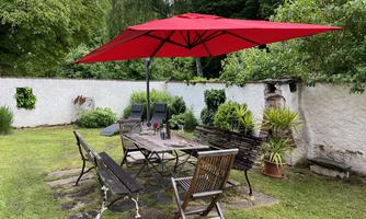 An inviting garden area with a large red sun umbrella. Surrounded by plants, there is a table with chairs, perfect for relaxing hours outdoors.