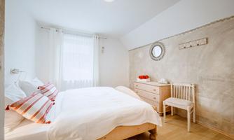A bright bedroom with a large bed and white bedding. The walls are plain, and there is a mirror as well as a dresser and a chair.