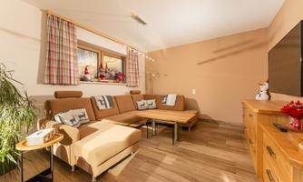 A cozy living room with a beige sofa and a wooden table. Curtains hang on the walls, and there are some decorative elements.