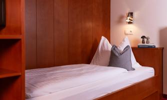 A cozy single bed in a wooden frame with pillows. In the background, a reading lamp and a nightstand can be seen.