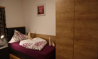 A cozy bedroom with a single bed and colorful cushions. A picture hangs on the wall and a wooden wardrobe stands next to the bed.