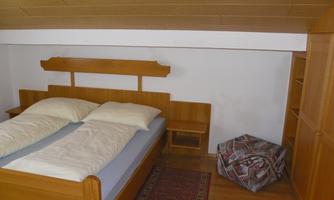 A cozy bedroom with a wooden bed and two pillows. Next to the bed is a small table and a padded stool.