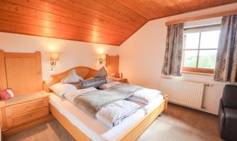 A cozy bedroom with a wooden bed and a warm roof. The walls are light, and large windows let in plenty of daylight.