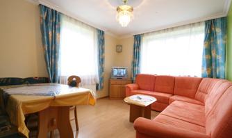 A cozy living room with an orange sofa and a table. Large windows let in plenty of light, and the room has colorful curtains.