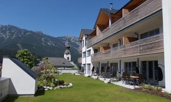 A modern building with balconies and a well-kept garden. In the background, mountains and an old church are visible.