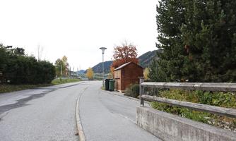 A quiet street with a wooden shed and trees on the side. In the background, hills and a gentle landscape can be seen.