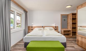 A modern bedroom with a double bed and a green stool. Bright colors and plenty of daylight create an inviting atmosphere.
