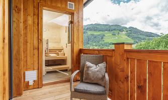 A cozy wooden balcony with a chair and a cushion. In the background, green hills and a clear sky are visible.