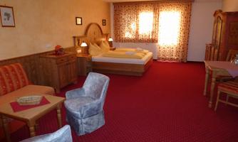 A cozy room with a double bed and rustic wooden furniture. The room features a seating area and large windows that let in plenty of light.