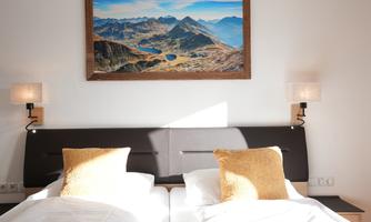A bright bedroom with two beds and golden pillows. Above the bed hangs a large landscape painting of the mountains.