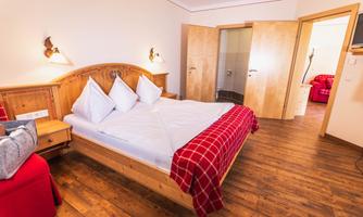 A cozy bedroom with a double bed and light wooden furniture. The room is warm and inviting, with red accents and natural light.