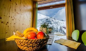 A fruit basket with apples and bananas is on a table. In the background, you can see snow-covered mountains through a window.