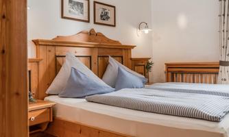 A cozy bedroom with a wooden bed and cushions in blue covers. Pictures hang on the wall, and there is bright lighting.