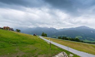 A green meadow with a winding path and scattered huts. The sky is cloudy and shows signs of impending rain.