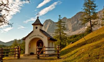 A small church stands in a picturesque mountain landscape. The surroundings are green and surrounded by trees, with majestic mountains in the background.