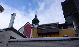 A view of a colorful building with a tall, pointed church tower in the background. The sky is partly cloudy and there is snow on the roofs.