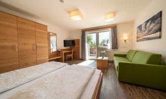 A bright hotel room with wooden furniture and a large window. There is a double bed, a table with chairs, and a green sofa.