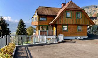 A charming wooden house with a large parking area and a beautiful view of the mountains. It is surrounded by trees and has a clear blue sky in the background.