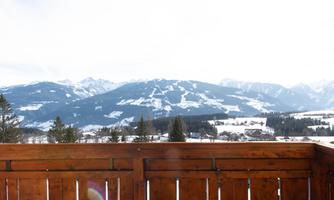 A majestic mountain landscape with snow-covered hills and a bright blue sky. In the foreground, a wooden railing is visible.