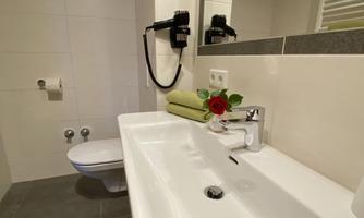 A modern bathroom with an elegant sink and a large mirror. On the shelf, there are green towels and a red rose.
