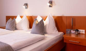 A cozy bedroom with two beds, white bedspreads, and gray pillows. On the wall, there are modern night lights mounted, and a wooden wardrobe stands beside it.
