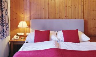 A cozy bedroom with a double bed and red pillows. Curtains hang on the wooden wall, and a bedside lamp provides light.