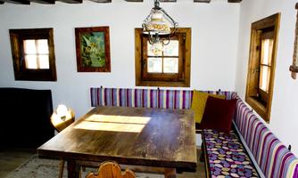 A cozy dining area with a large wooden table and a colorful bench. Bright walls and windows create an inviting atmosphere.