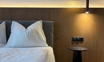 A modern bedroom with a large bed and white pillows. A stylized bird graphic hangs on the wall, and there is a small table as well as a cozy lamp.