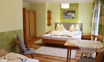 A cozy bedroom with two beds and a table. The walls are painted in soft shades of green, and there is a bright window decoration.
