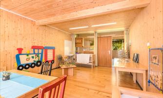 A bright room with wooden walls and a colorful toy train on the wall. There are several tables and chairs as well as a small kitchenette in the background.