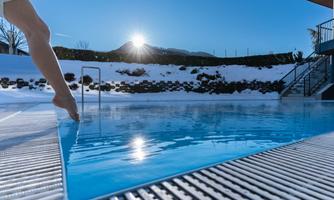 A foot approaches a clear pool in the snow. The sun shines over the mountains.