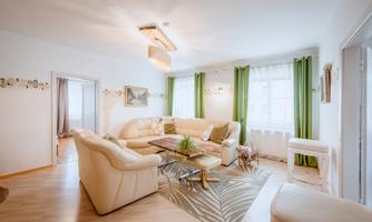 A bright living room with comfortable, white furniture and green curtains. The room is friendly and inviting, with a small table and decorative plants.