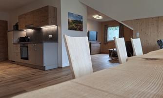 A bright, modern kitchen with wooden shelves and a dining table in the foreground. In the background, there is a living room with windows and a television visible.