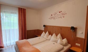 A cozy bedroom with a large bed and white pillows. The room has a modern chandelier and a view of the garden through a glass door.