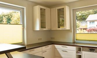 A modern kitchen with white cabinets and a bright table. The windows are fitted with yellow blinds and offer a view outside.