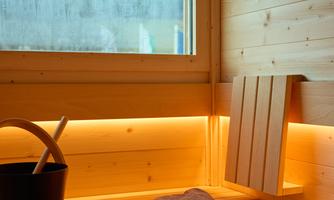 A cozy sauna with wooden paneling and soft lighting. On the bench, there is a towel and a bucket.