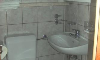 A small bathroom with ceramic tiles. There is a toilet, a sink, and towels on a shelf.