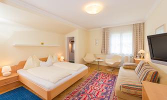 A cozy room with a double bed and a carpet. The walls are painted in soft colors and there is a sitting area with a TV.