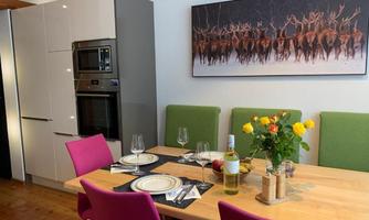 A modern dining area with a wooden table and colorful chairs. A picture of deer in a winter landscape hangs on the wall.