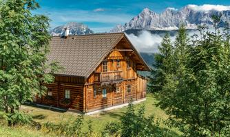 A cozy wooden house surrounded by trees and green grass. In the background, majestic mountain peaks and a clear sky can be seen.