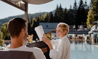 A relaxed atmosphere by the pool with a smiling staff member showing something to a guest. Trees and mountains are visible in the background.